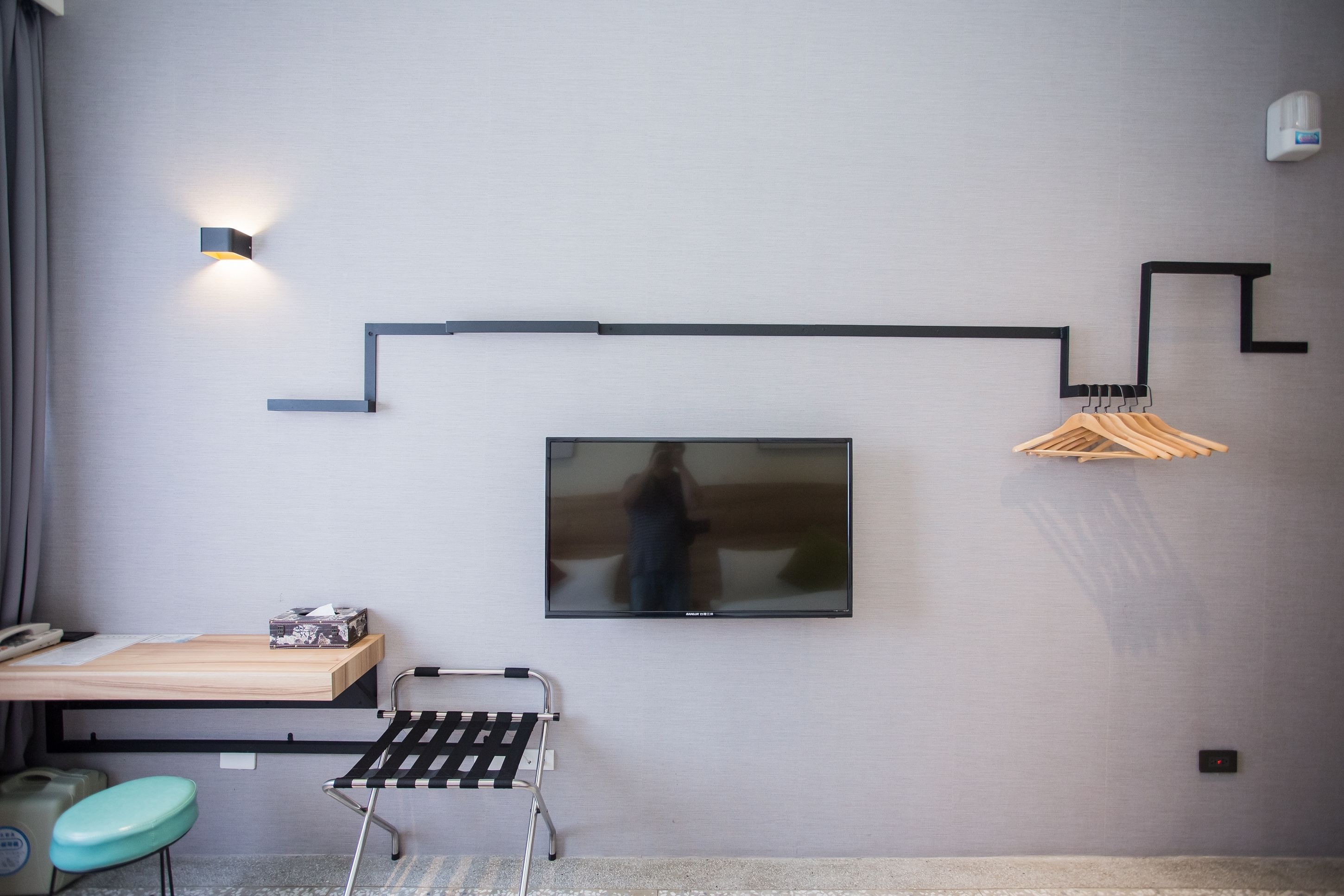 Una habitación moderna con un televisor de pantalla plana montado en la pared, un escritorio de madera con un teléfono, un taburete pequeño y una silla de metal negro.