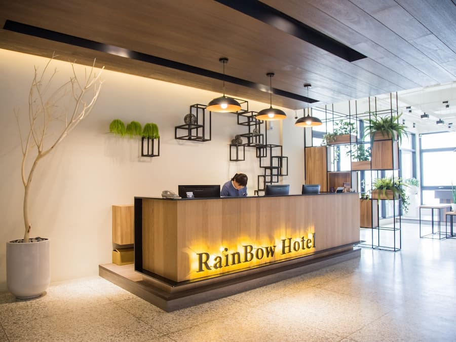 A reception desk at the Rainbow Hotel with a modern design, including a wooden reception area, pendant lighting, and potted plants.