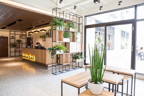 Zona de recepción de un hotel con un mostrador de madera, una exhibición de plantas y vistas a la calle.