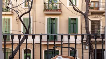 Un balcon avec un plateau en bois sur lequel reposent deux tasses blanches, un arbre au premier plan et un bâtiment avec des volets verts et des balcons en arrière-plan.