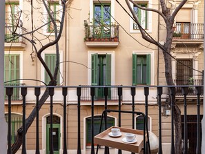 A balcony with a wooden tray holding two white cups, a tree in the foreground, and a building with green shutters and balconies in the background.