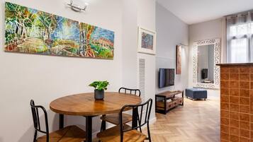 Un salon moderne avec une table de salle à manger ronde en bois, des chaises en métal noir, une plante en pot, un téléviseur à écran plat, un miroir décoratif et un parquet à motifs en chevrons.