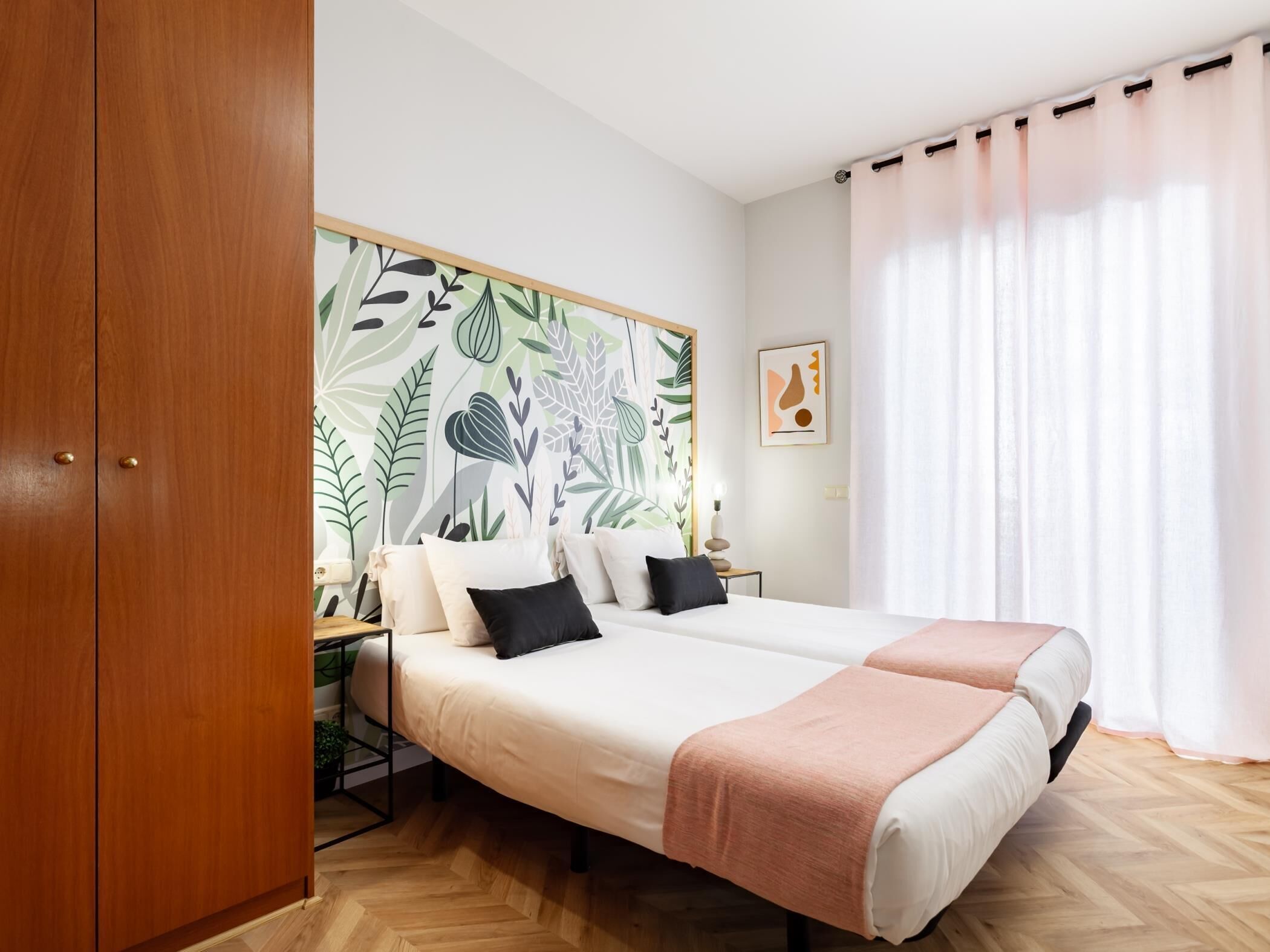 Une chambre avec un grand lit, une armoire en bois, un mur orné d’un motif de feuilles, une table de chevet avec une lampe et une fenêtre avec des rideaux.