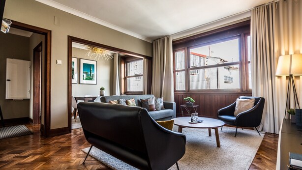 Un salon comprenant un canapé, un fauteuil et une table basse, de grandes fenêtres avec des rideaux et une lampe.