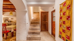 Una escalera que conduce a una puerta de madera, un comedor con una mesa y sillas, y un gran tapiz decorativo en la pared.