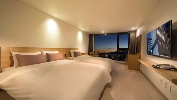 A hotel room with two beds, a view of the cityscape, and a large window. - Pino Hotel (Seogwipo)