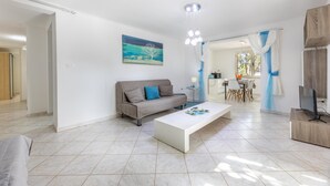 Een moderne woonkamer met een bank, salontafel en televisie. Op de muur hangt een schilderij en er is een eethoek op de achtergrond.