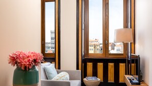 A cozy room with a sofa, a small table with a vase of flowers, a lamp, and a view of buildings through large windows. - Authentic Studio in Bolhão Market (Oporto)