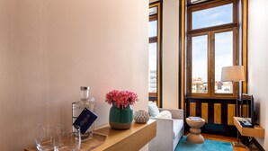 A hotel room with a wooden counter, a bottle of liquor, two glasses, a vase with flowers, a sofa, a lamp, and a view of buildings through large windows. - Authentic Studio in Bolhão Market (Oporto)