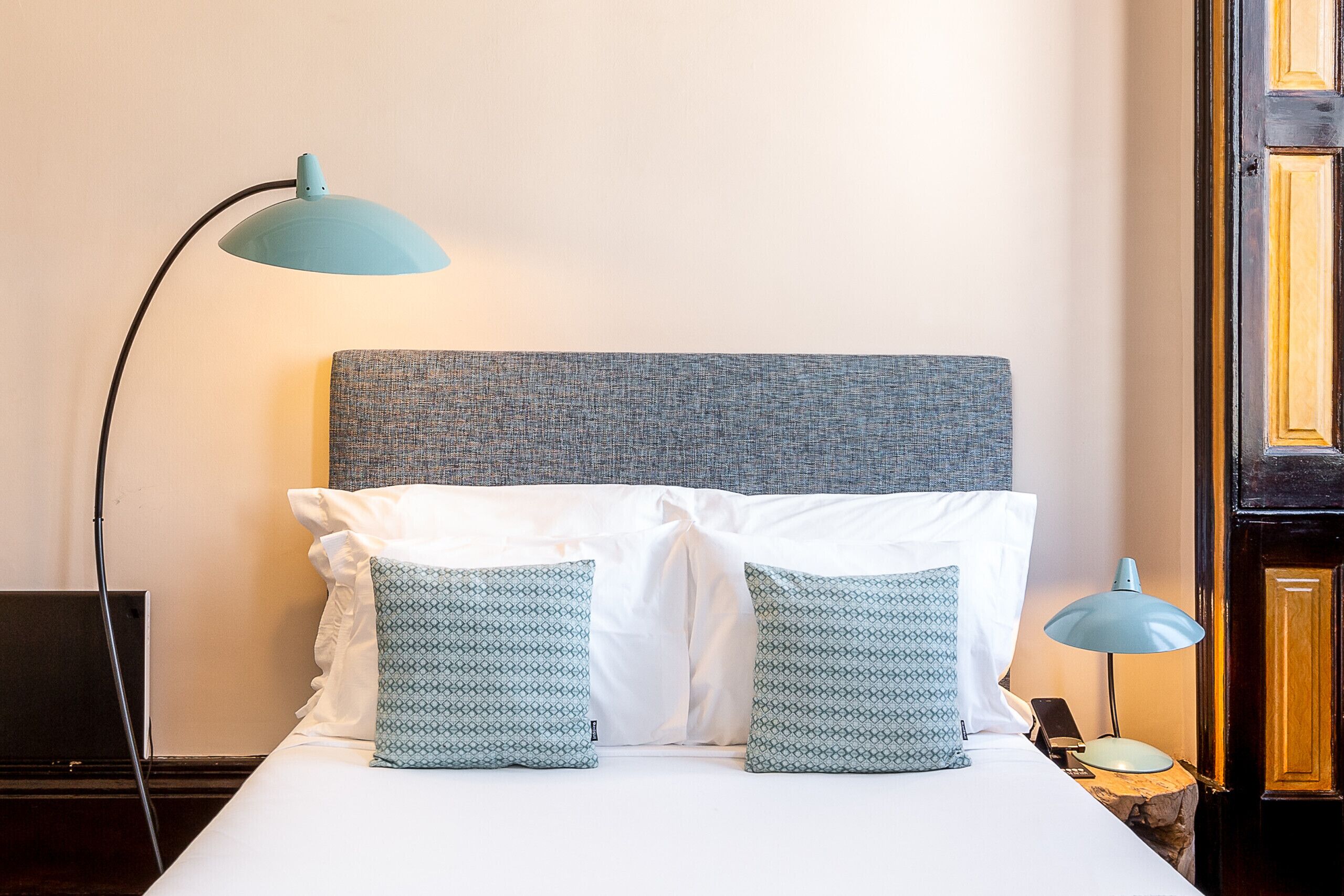 Un lit bien fait, avec des oreillers blancs et à motifs, une tête de lit grise et deux lampes bleues de chaque côté.
