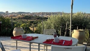 An outdoor patio with a table set for two, a large umbrella, and a view of a cityscape in the background. -  "Little cottage
 surrounded by olive and almond trees (Noto)