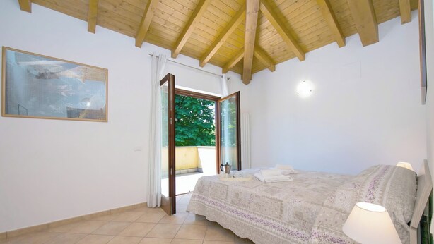 Une chambre à coucher avec un lit, une table de chevet, une lampe, un tableau, une fenêtre et une vue sur la verdure à l’extérieur.