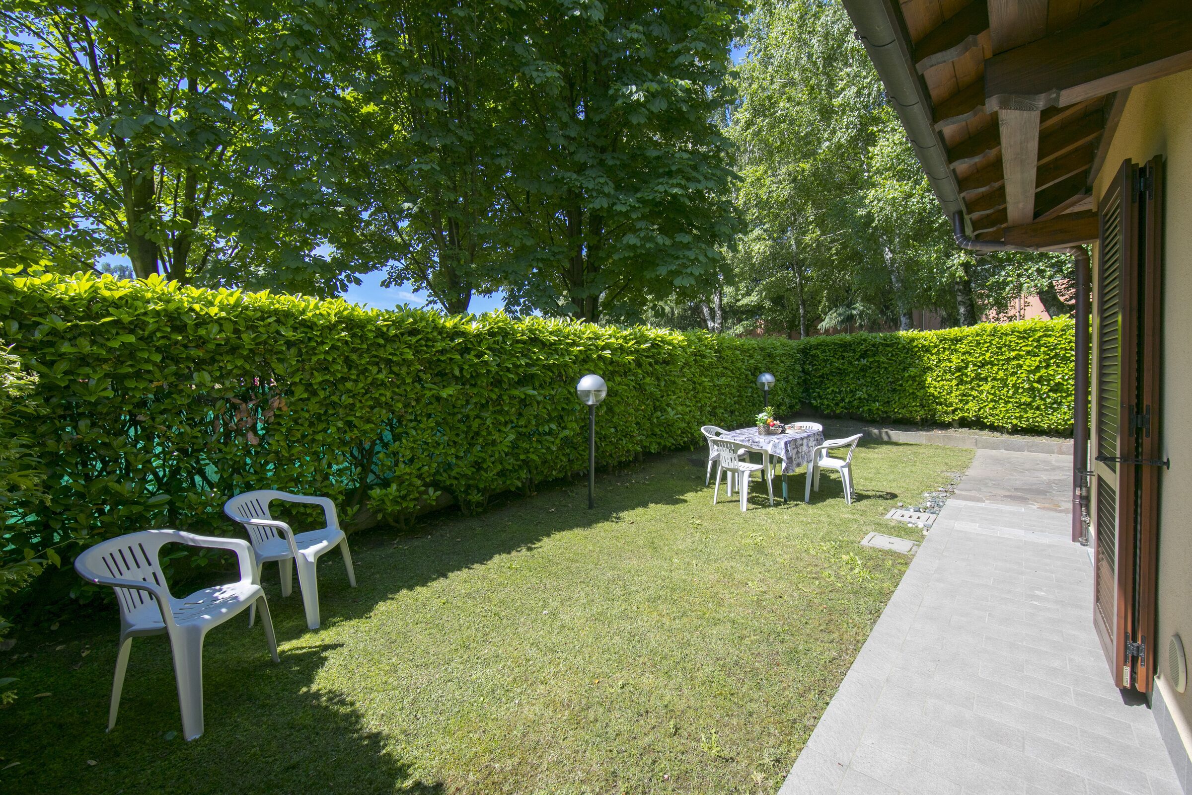 Ein gepflegter Garten mit einer Terrassenfläche, inklusive Tisch und Stühlen, umgeben von einer Hecke und Bäumen.