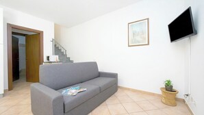 Un salon avec un canapé gris, un tableau encadré au mur, un téléviseur fixé au mur et une petite plante posée sur une table d’appoint.