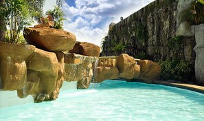 Piscina com água cristalina, rodeada por pedras grandes e um penhasco rochoso.