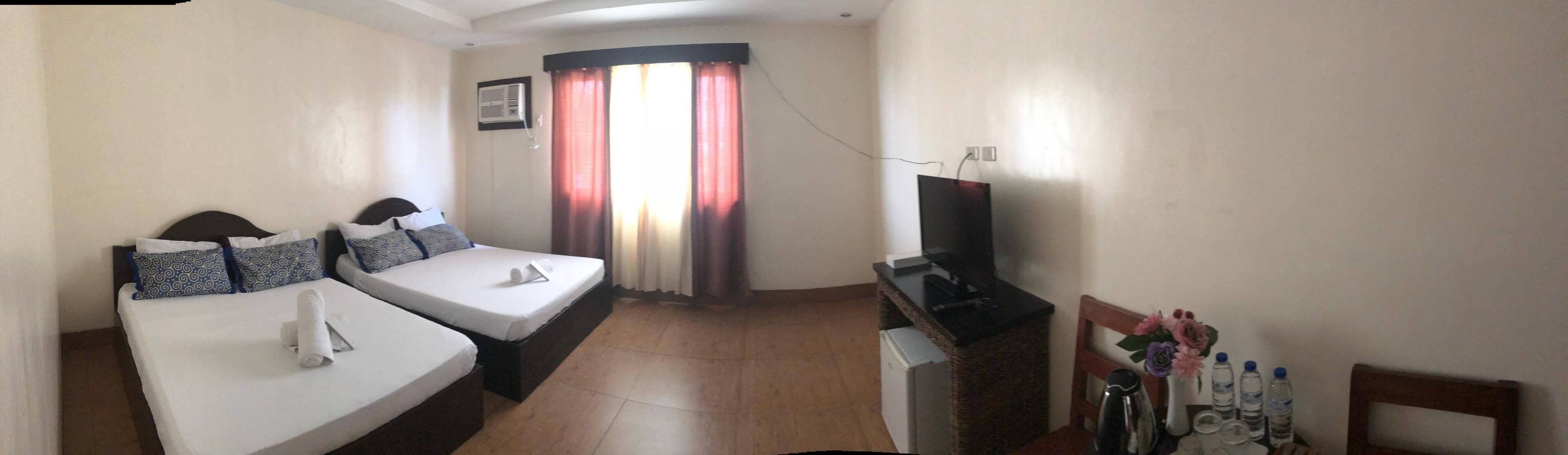 A hotel room with two beds, a TV, a desk with a computer, a window with curtains, and a wall-mounted air conditioner.
