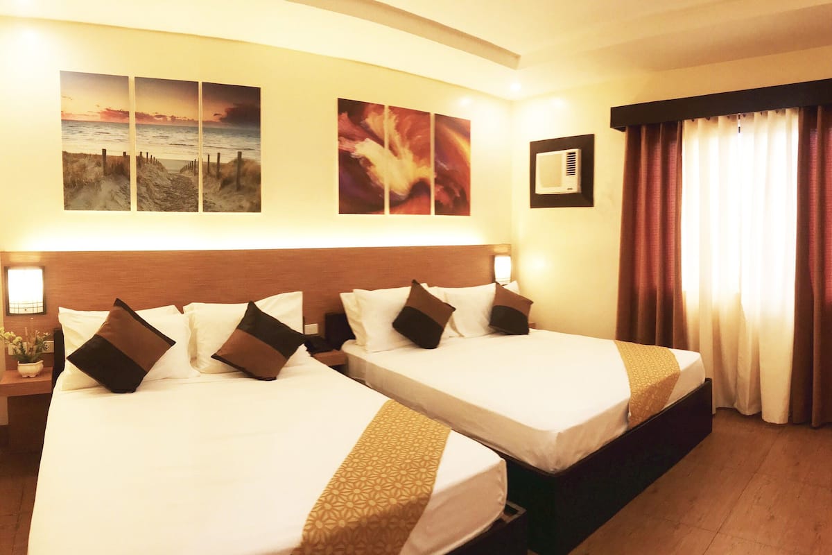 Quarto de hotel com duas camas, mesa de cabeceira com abajur, ar condicionado fixado na parede e janela com cortinas.