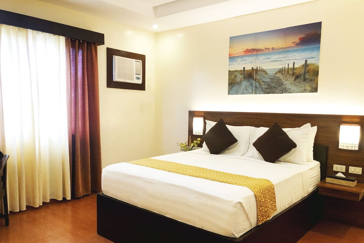 Quarto de hotel com uma cama grande, mesa de cabeceira, ar condicionado fixado na parede e um quadro com uma paisagem de praia.