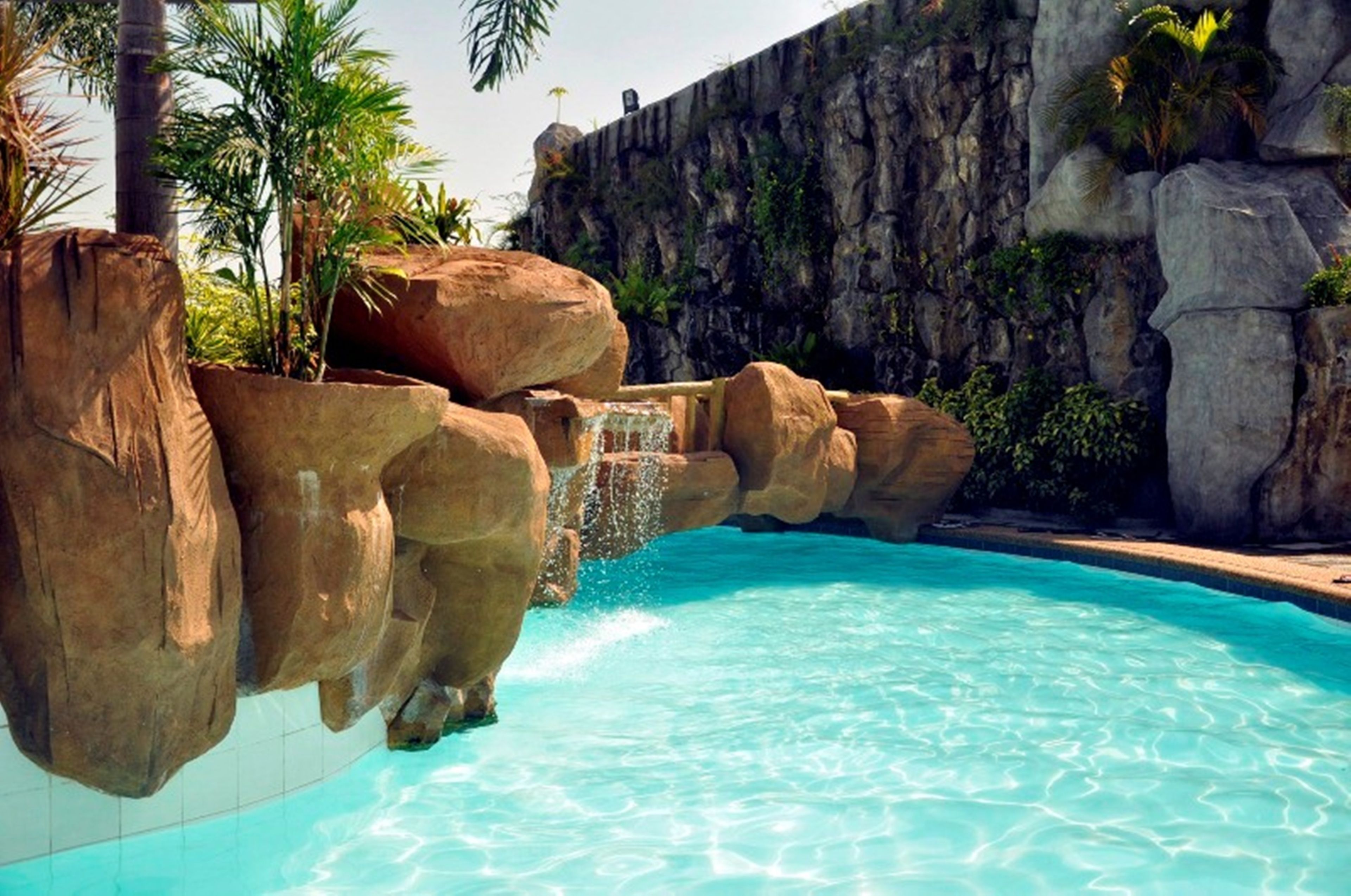 A pool with clear blue water, surrounded by large rock formations and palm trees.