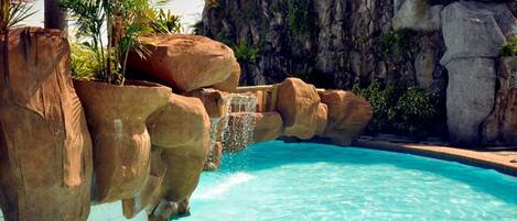 A pool with clear blue water, surrounded by large rock formations and palm trees.