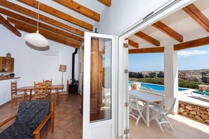 A modern room with wooden beams, a dining area, and a view of a pool and rocky landscape. - Sea Frontline House With 180 Degree Panoramic Sea Views (Es Mercadal)