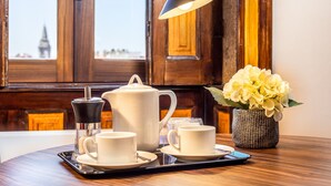 Una colazione allestita con una macchina del caffè, un teiera, tazze e piattini su un vassoio, un vaso di fiori e una lampada su un tavolo vicino a una finestra con vista su un edificio.