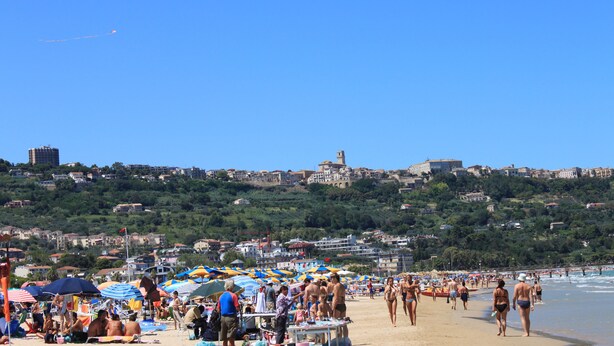 Una playa concurrida, gente tomando el sol y caminando, con un paisaje urbano al fondo.