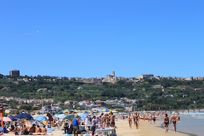 Una playa concurrida, gente tomando el sol y caminando, con un paisaje urbano al fondo.
