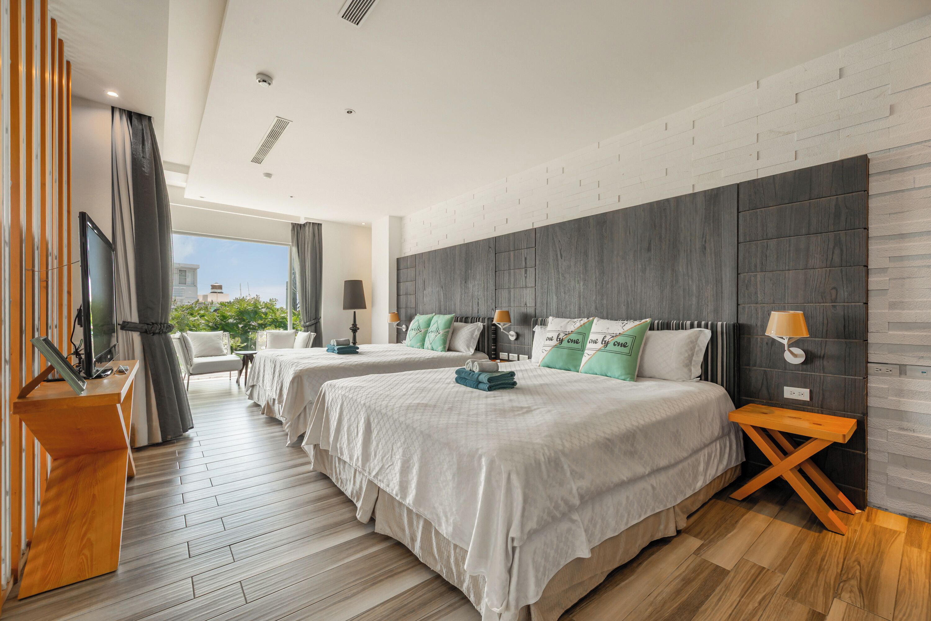 A hotel room with two beds, a TV, a desk, and a view of greenery outside.