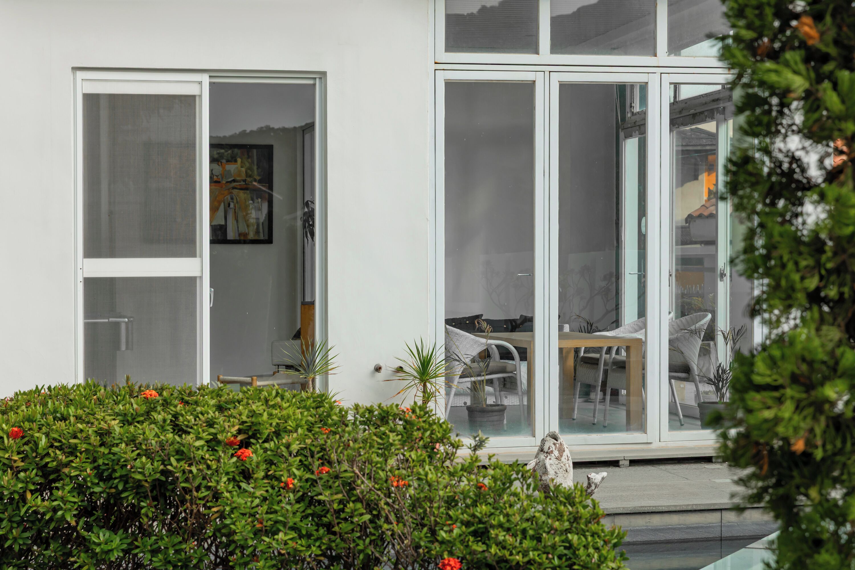 Ein modernes Gebäude mit großen Glasfenstern und -türen, ein sichtbarer Innenbereich mit Essbereich sowie ein gepflegter Garten mit roten Blüten.