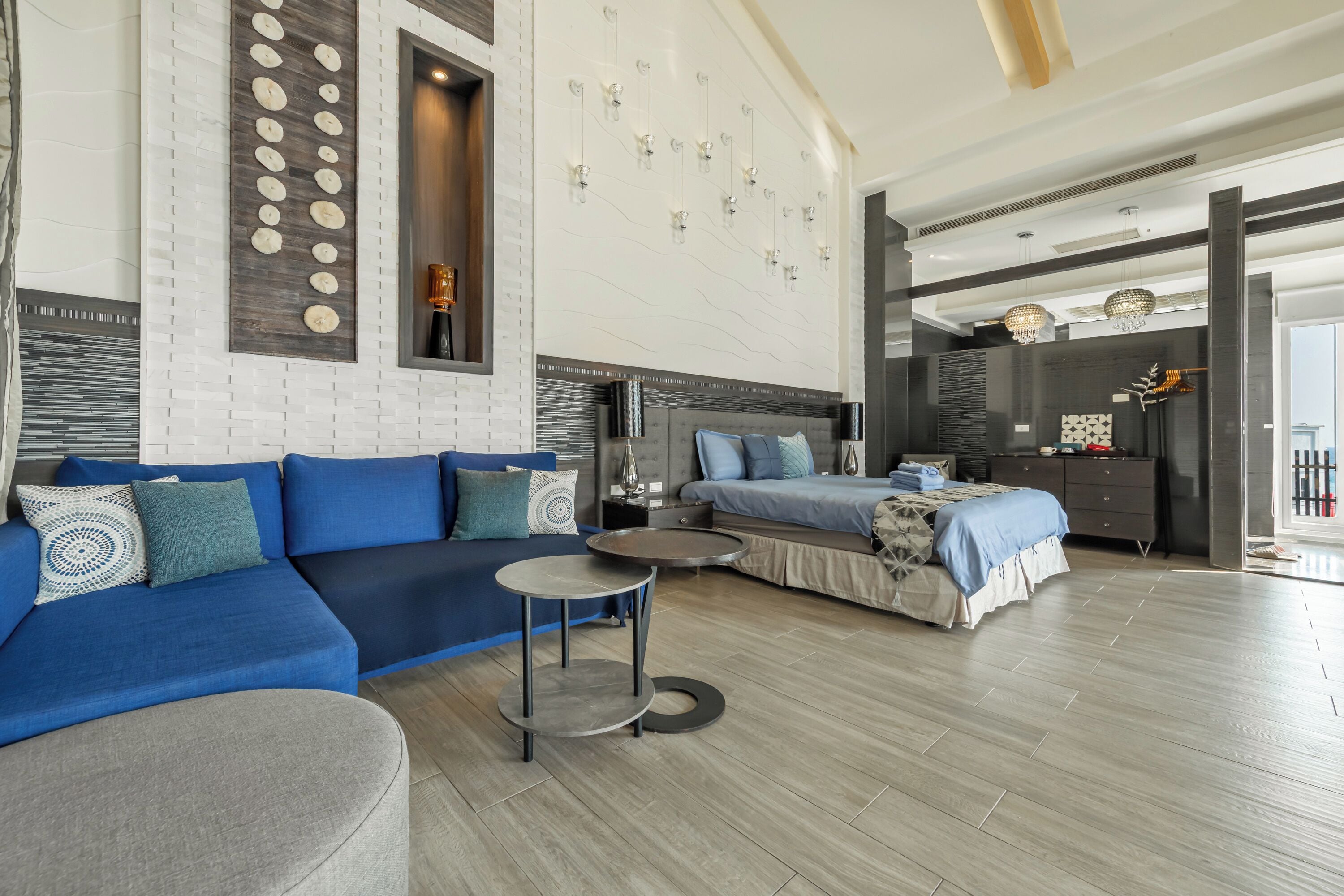 Una habitación de hotel moderna con sofá modular, una mesita redonda, cama con colcha estampada y una pared decorativa con luces colgantes.