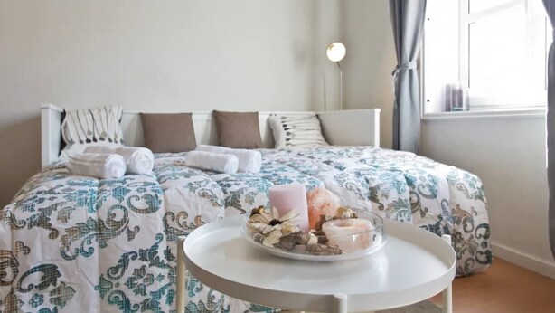 Un letto rifatto con cura, una coperta a motivi decorativi, un comodino con una candela e degli snack, e una finestra con tende.