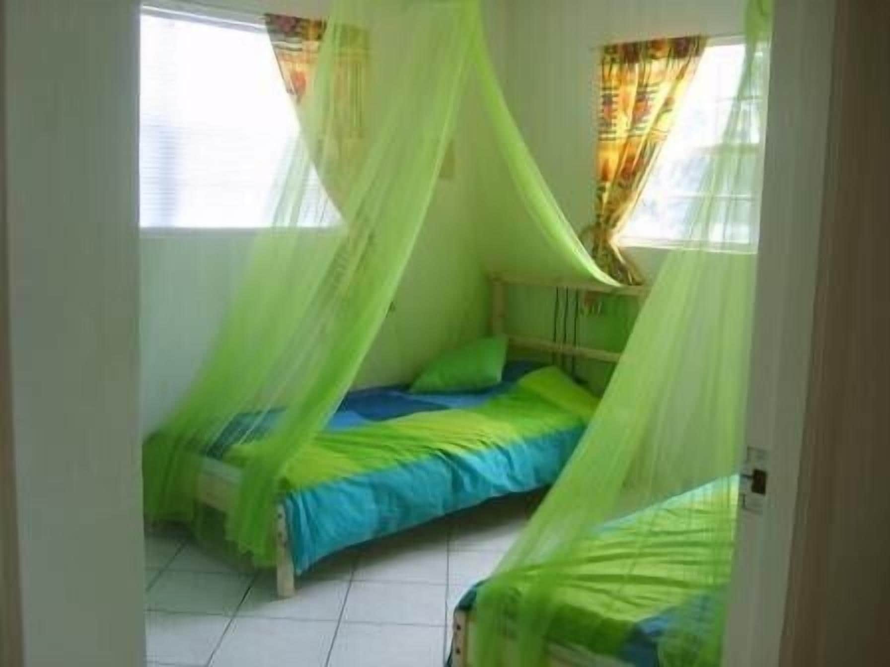 Una habitación con dos camas, cada una con ropa de cama verde y azul. Hay ventanas con cortinas, y el suelo está cubierto de baldosas.