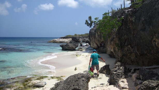 Een strand met helder turquoise water, rotsformaties en een persoon die over het zand loopt.
