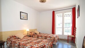Une chambre d’hôtel avec deux lits, une fenêtre avec des rideaux rouges, un tableau encadré au mur et une petite table avec une lampe.