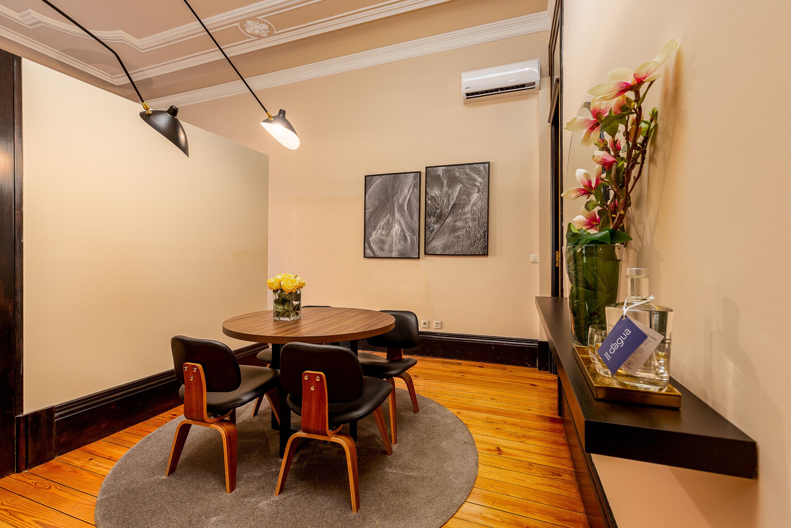 A dining area with a round table, four chairs, a vase with flowers, and two framed artworks on the wall.