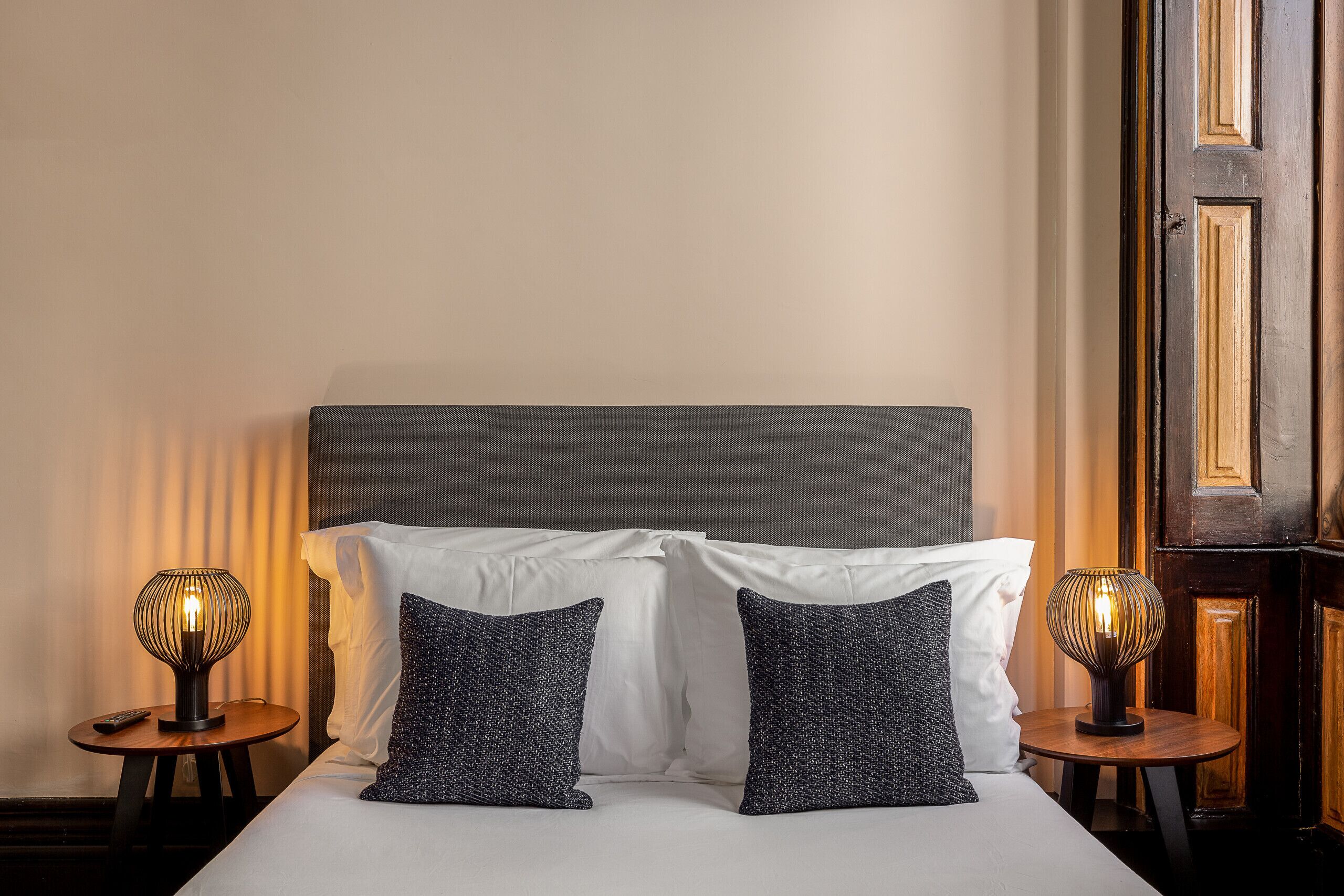 A neatly made bed with white linens and two dark patterned pillows, flanked by two small tables with lamps, one on each side.