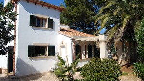 A two-story white building with a tiled roof, green shutters, and a small garden with palm trees and shrubs. - Lovely holiday villa. Pool and beach (Cala Serena)