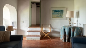 A room with a brick floor, a wooden-framed armchair, a table with a floral tablecloth, a lamp, and a framed painting on the wall. - House with pool. Idyllic setting in hills near Lucca and coast WiFi Aircon       (Torcigliano)