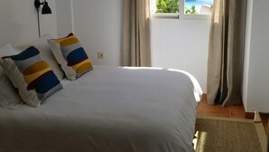 Une chambre avec un grand lit, une fenêtre avec des rideaux, une lampe fixée au mur et un tableau encadré.
