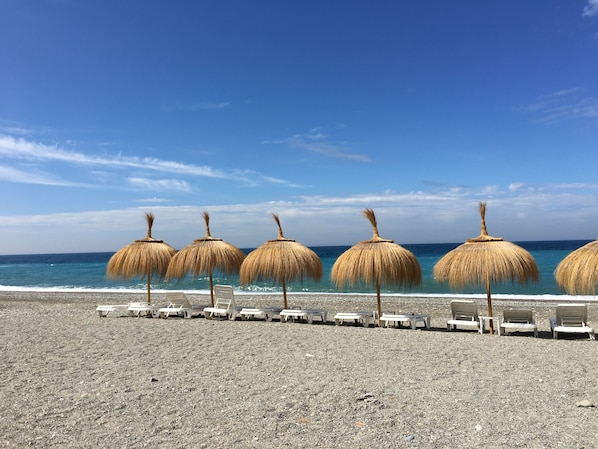 A beach with thatched umbrellas and white lounge chairs aligned along the shore. - CASA EL MERO LA HERRADURA (Almuñécar)