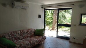 Un salon avec un canapé à motifs, une porte-fenêtre coulissante donnant sur un jardin et un climatiseur fixé au mur.