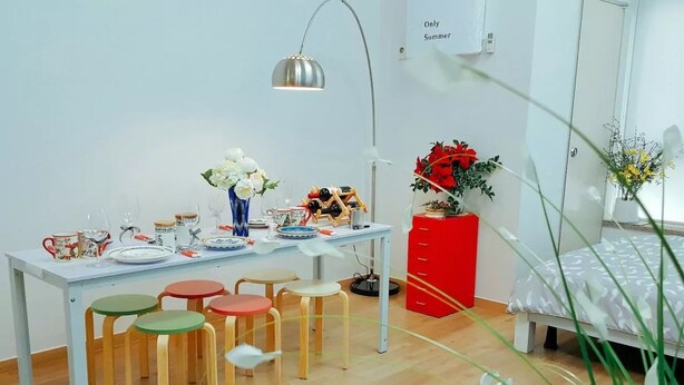 A small, bright room with a wooden floor, a white table with colorful stools, a red cabinet, and a bed with a white and gray patterned bedspread.
