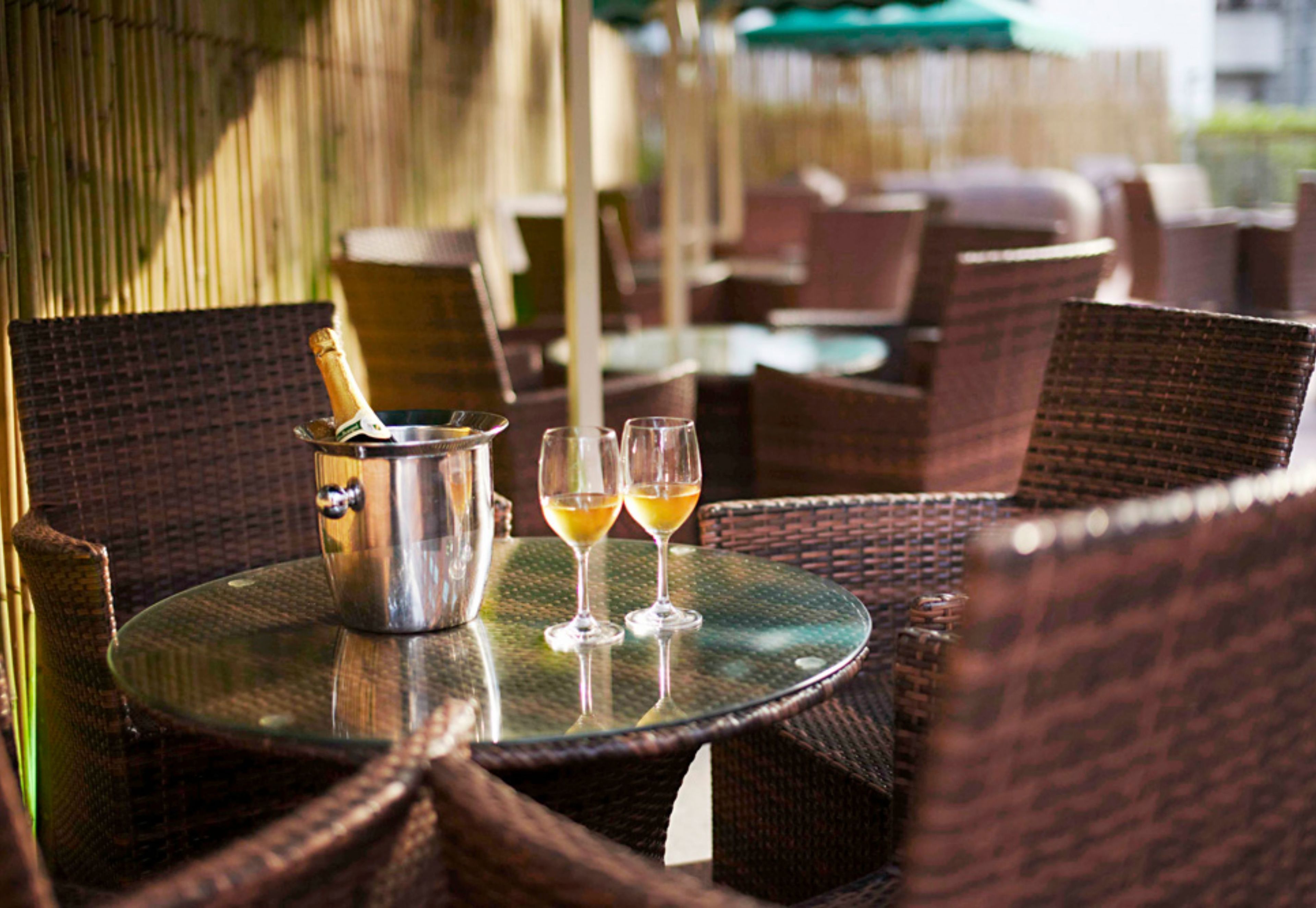 A round table with wicker chairs, a metal bucket with a bottle, and two glasses of wine.
