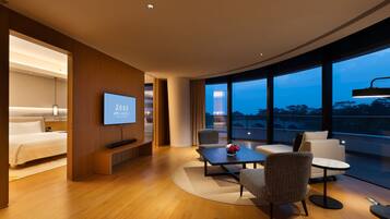 A modern hotel room with a large bed, a TV, a seating area with a coffee table, and a view of the city at dusk.