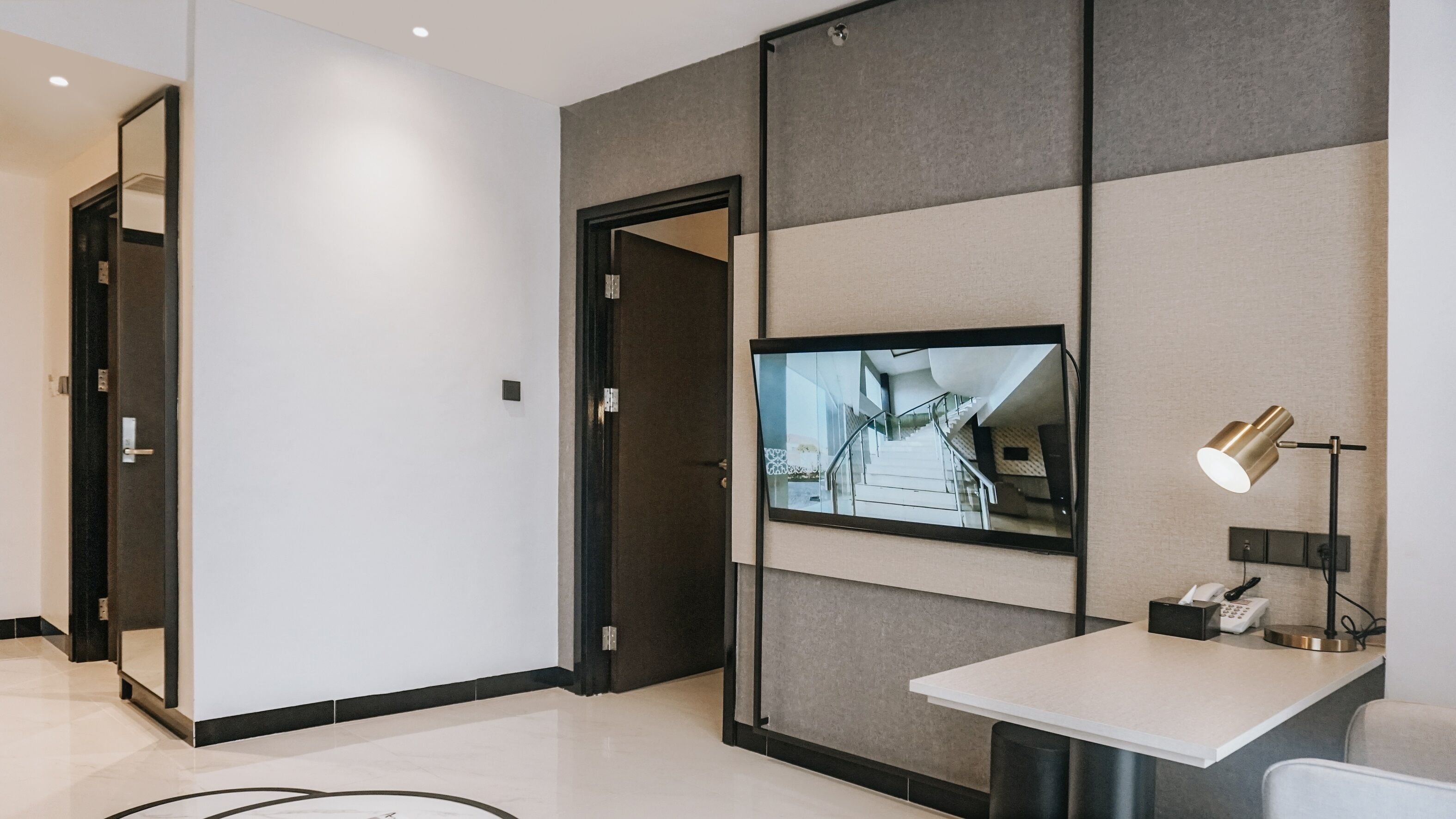 Una camera d'hotel moderna con una grande TV a schermo piatto montata su una parete strutturata, una scrivania con una lampada e un telefono.