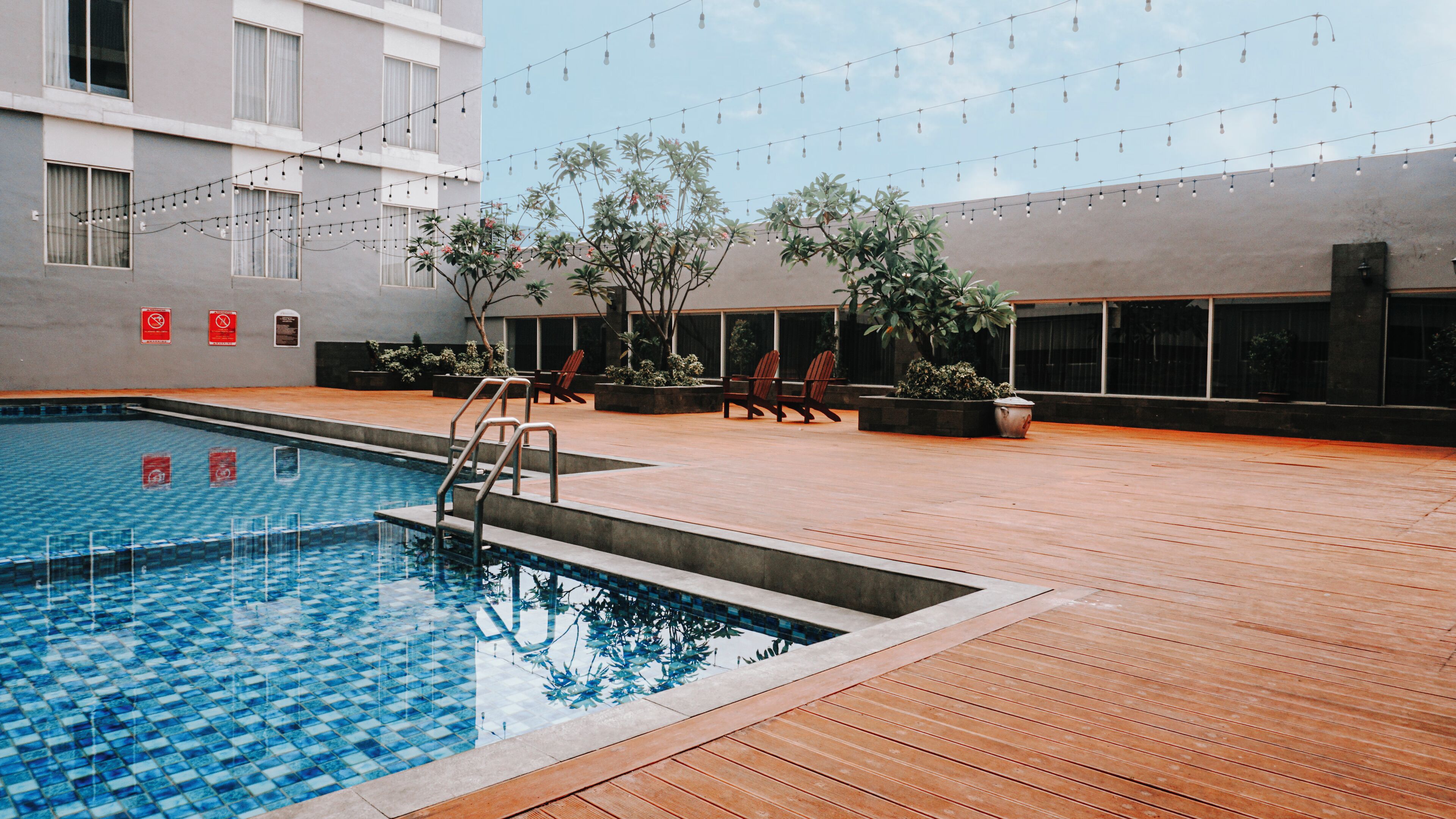 A swimming pool with a wooden deck, surrounded by potted plants and outdoor seating.