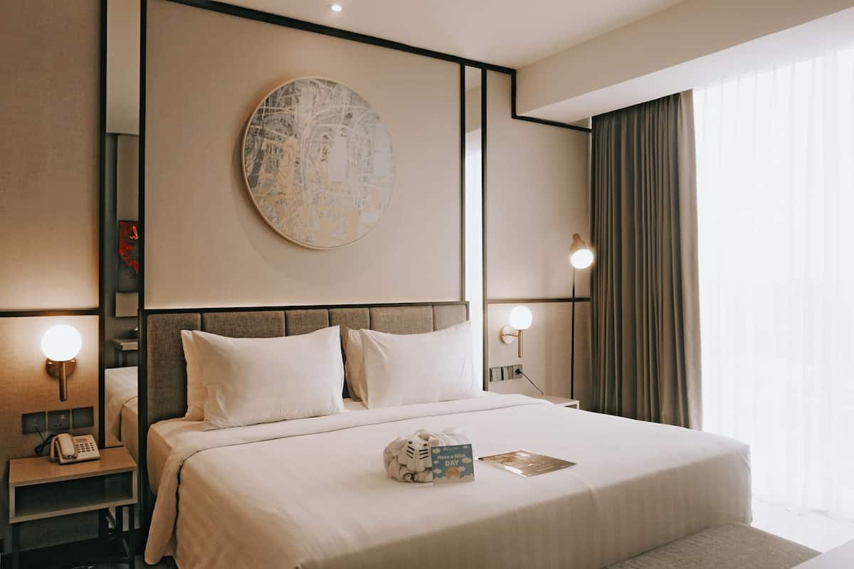 A modern hotel room with a large bed, bedside lamps, a round artwork on the wall, and a view of the outdoors through sheer curtains.