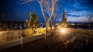 A rooftop terrace with a view of the city at night, featuring a metal bench, a table, and a small garden area with trees. - Hotel AMARE (Suwon)