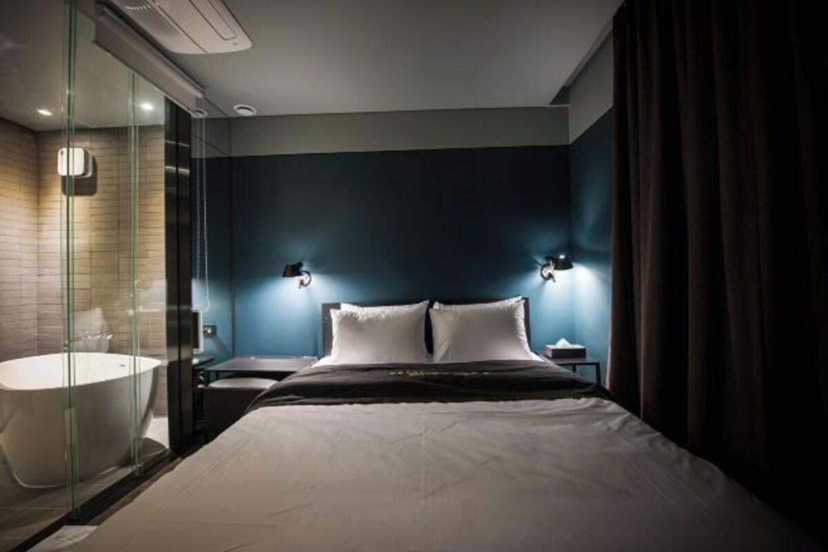 Habitación de hotel moderna con una cama grande, cabina de ducha de vidrio y una mesita de noche con lámpara.